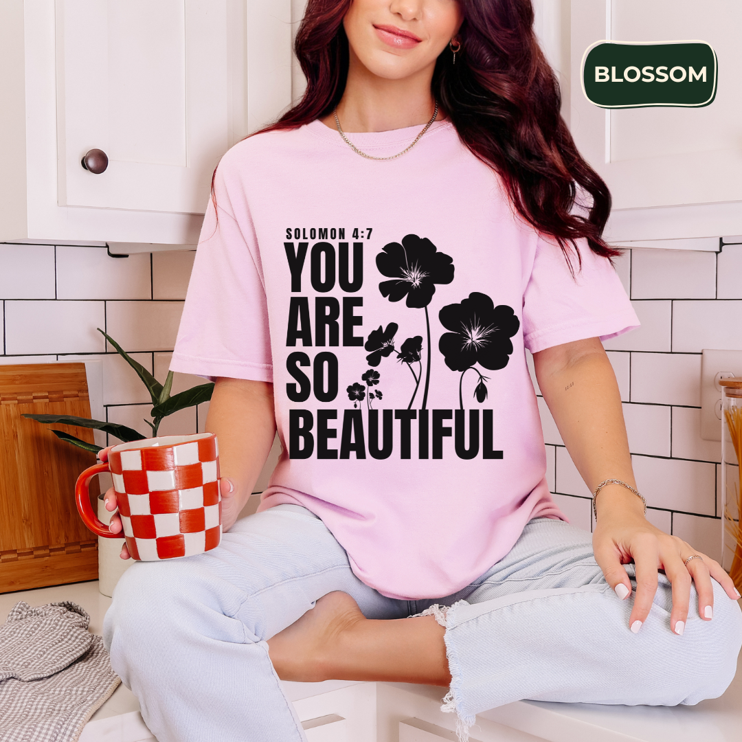 Woman wearing a pink t-shirt with floral design and text, holding a mug in a kitchen.
