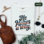 White t-shirt with 'He Makes a Way' text, brown bag, necklace, and plant on a light background