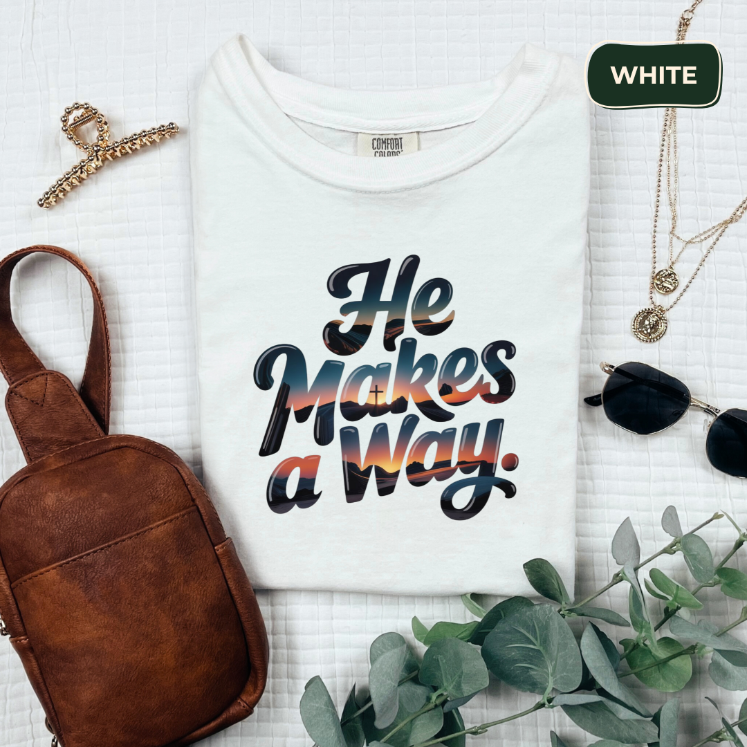 White t-shirt with 'He Makes a Way' text, brown bag, necklace, and plant on a light background